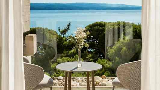 En borddekorasjon med blomster ved et bord foran vinduer med utsikt over havet og vegetasjon. (AI-generert tekst)