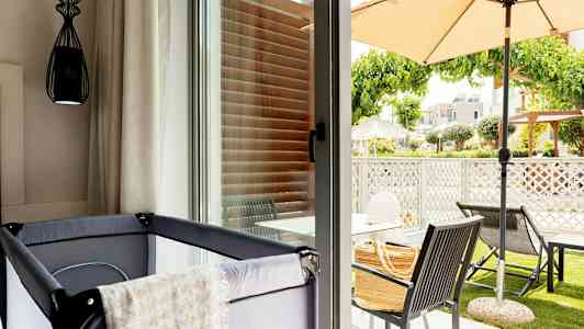 Bilde av et rom med barneseng nær et glassvindu som vender mot en terrasse med utemøbler og parasoll. (AI-generert tekst)