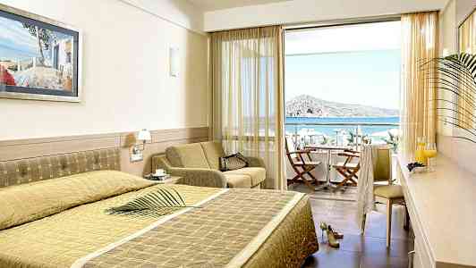 Hotellrom med seng, sitteområde og utsikt mot havet gjennom en balkong med stoler og bord. (AI-generert tekst)