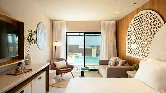 Et elegant hotellrom med moderne møbler, en sofa, en lenestol, et sengområde med dekorativt hodegjerde, et stort speil og glassdører som åpner ut mot et svømmebasseng utendørs. (AI-generert tekst)