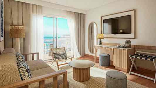 Et lyst og moderne hotellrom med utsikt over havet. Det er en sofa, et sofabord, en stol og en TV på veggen. (AI-generert tekst)