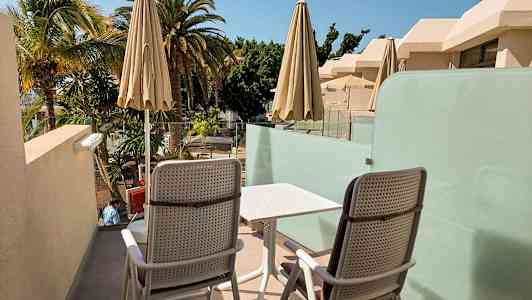 To stoler og et bord på en terrasse med utsikt over palmer og bygninger. (AI-generert tekst)