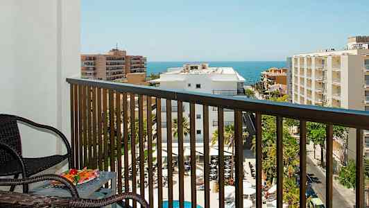 Utsikt fra en balkong med møbler over et basseng, bygninger og havet i bakgrunnen. (AI-generert tekst)