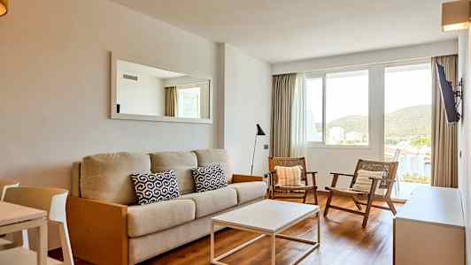 En stue med en beige sofa, to lenestoler og et hvitt sofabord. Det er et vindu med utsikt til grønt. (AI-generert tekst)