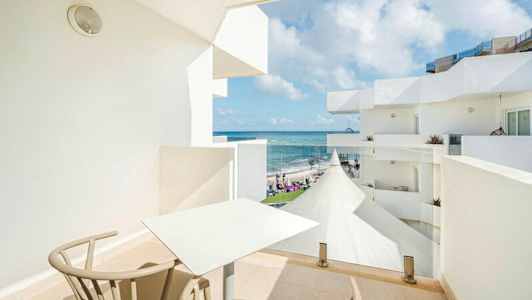 En balkong med bord og stoler, med utsikt over havet og hvite bygninger. (AI-generert tekst)