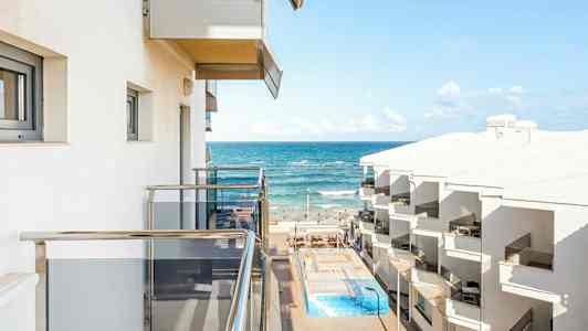 Utsikt fra balkong mot havet med hotellbygninger og basseng. (AI-generert tekst)