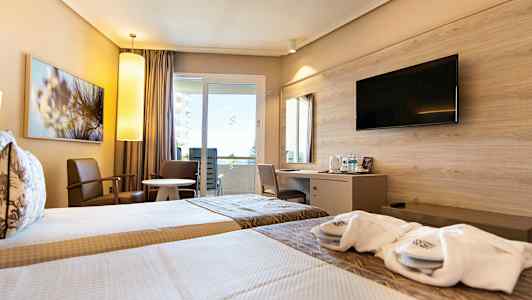 Et hotellrom med to enkeltsenger, sittemøbler ved et rundt bord samt en skrivebordskrok med en veggmontert TV over. (AI-generert tekst)