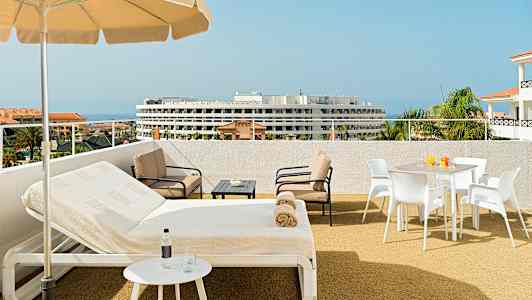 Utendørs terrasse med parasoll, solsenger og sitteområde. Havet og en bygning er synlige i bakgrunnen. (AI-generert tekst)