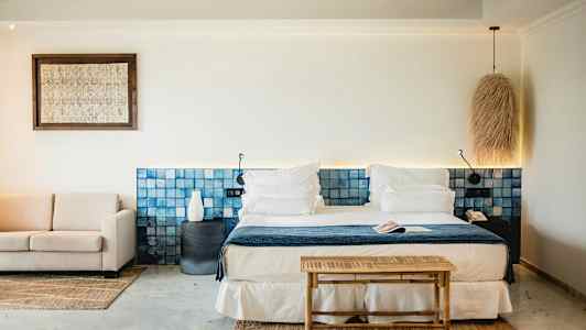 Et stilrent hotellrom med en dobbeltseng, blå hodegavl, sittegruppe og lyse dekorative elementer. (AI-generert tekst)