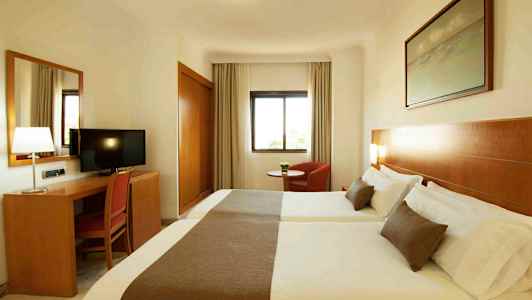 Et hotellrom med to senger, et skrivebord med en stol og en TV, samt et vindu og en lenestol. (AI-generert tekst)