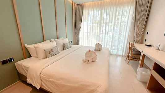 Bilde av et moderne hotellrom med en stor oppredd seng, hvitt sengetøy og dekorative puter, samt håndklær formet som svaner. Rommet har vinduer med lyse gardiner og en arbeidsplass ved siden av. (AI-generert tekst)