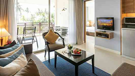 Moderne innredet stue med sofa, lenestol, glassbord med frukt, TV og kjøkkenkrok, samt balkong med utsikt mot palmer. (AI-generert tekst)