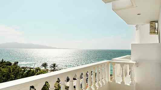 Utsikt fra en balkong mot havet og en klar himmel, med palmer og trekroner nedenfor. (AI-generert tekst)