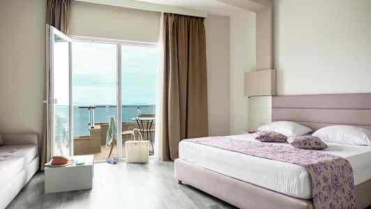 Et hotellrom med en stor seng, glassdører ut til en balkong og utsikt over havet. (AI-generert tekst)