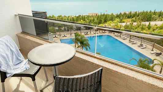 Utsikt fra en balkong med bord og stoler som viser et basseng og havet i bakgrunnen. (AI-generert tekst)