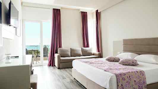 Et hotellrom med en dobbeltseng, en sofa og en balkong med utsikt mot havet. (AI-generert tekst)