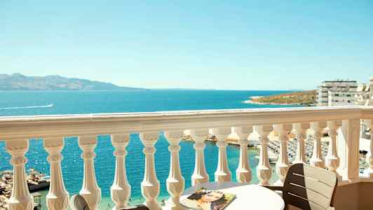 Utsikt fra en balkong med et hvitt rekkverk over havet, et bord og en stol. (AI-generert tekst)