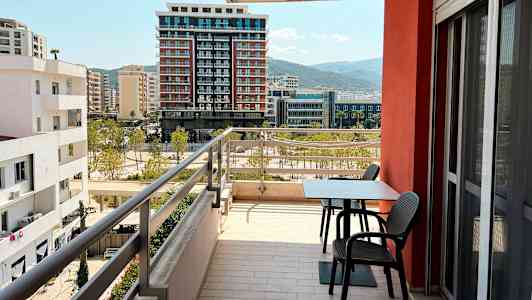 En balkong med bord og stoler med utsikt over omkringliggende bygninger og fjell i det fjerne. (AI-generert tekst)