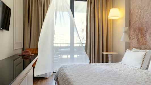 Et hotellrom med en seng, TV, bord og stol, samt et vindu med hvite gardiner. (AI-generert tekst)