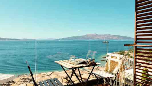 Utsikt fra en balkong mot havet og en strand med parasoller. På balkongen finnes et lite bord og en stol. (AI-generert tekst)