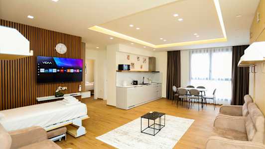 Et moderne hotellrom med åpen planløsning som inkluderer en sovesofa, TV, kjøkkenområde, spisebord og store vinduer med gardiner. (AI-generert tekst)