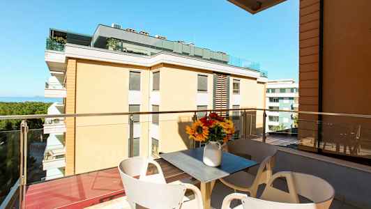 En balkong med utsikt over bygninger og havet. På balkongen er det et bord med en vase med blomster. (AI-generert tekst)