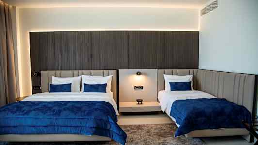 Bilde av et hotellrom med to senger kledd i blå sengtrekk og hvite puter. Fargerike tepper kan sees på gulvet. (AI-generert tekst)