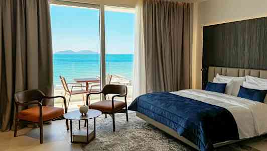 Et hotellrom med havutsikt, med en dobbeltseng, to stoler, et lite bord og en glassdør til en terrasse. (AI-generert tekst)