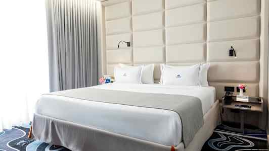 Et soverom med en stor seng, hvite laken og grått overkast. Bak sengen er det en polstret sengegavl, og på sidene står nattbord med lamper. (AI-generert tekst)