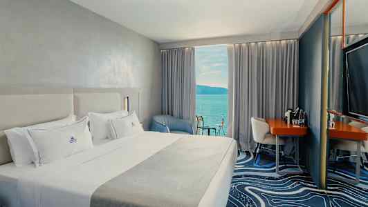 Et hotellrom med en stor seng, en lenestol og et skrivebord. Utsikt over havet fra et vindu med gardiner. (AI-generert tekst)