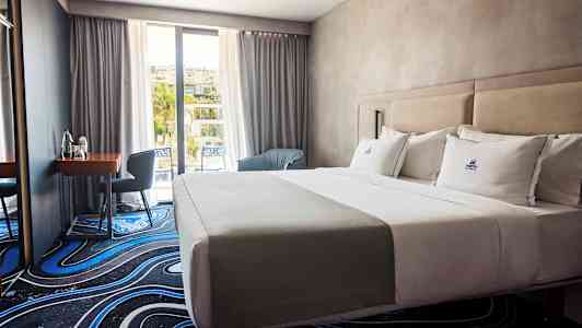 Et hotellrom med en oppredd dobbeltseng, et skrivebord med stol og vindu mot balkong. (AI-generert tekst)