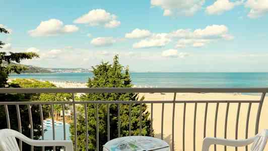 Utsikt fra en balkong mot en strand og havet, med stoler og et bord i forgrunnen. (AI-generert tekst)