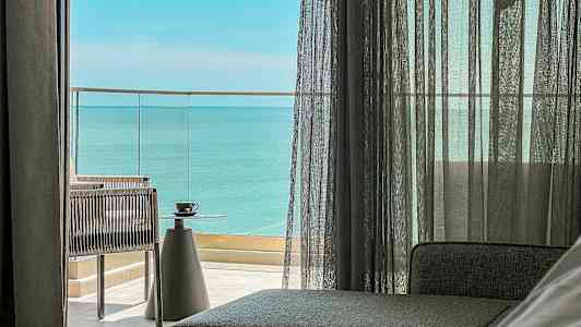 Utsikt fra et rom med glassdører som leder ut til en balkong med havutsikt. En kopp står på et bord. (AI-generert tekst)