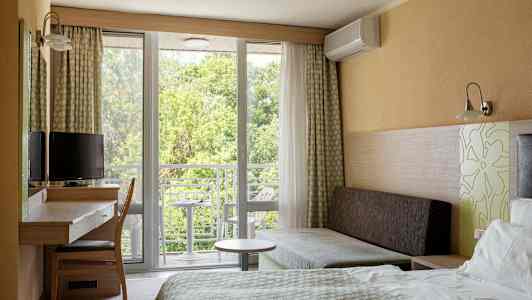 Et hotellrom med seng, skrivebord, stol og sofa. Vindu og balkong med utsikt mot grøntområder. (AI-generert tekst)