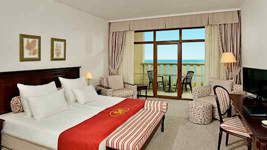 Et hotellrom med dobbeltseng, sittegruppe, et skrivebord og balkong med havutsikt. (AI-generert tekst)