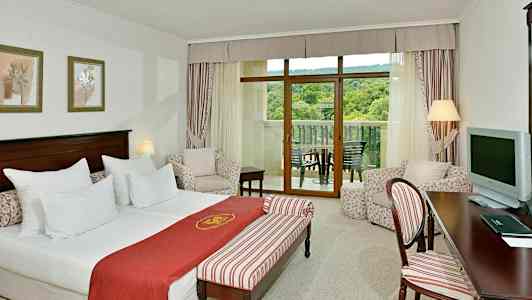 Hotellrom med seng, sittegruppe og balkong med utsikt over grøntområder. (AI-generert tekst)