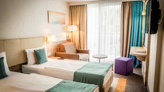 To enkeltsenger med turkise sengetepp i et hotellrom med vindu og sittegruppe. (AI-generert tekst)