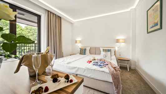 Hotellrom med dobbeltseng, nattbord og balkong med utsikt mot trær. Et bord med champagnekjøler og dessert. (AI-generert tekst)
