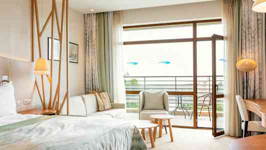 Et hotellrom med seng, sittegruppe, bord og åpen balkongdør. (AI-generert tekst)