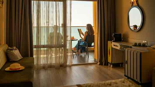 Hotellrom med en balkong der noen sitter og leser med utsikt over havet. (AI-generert tekst)