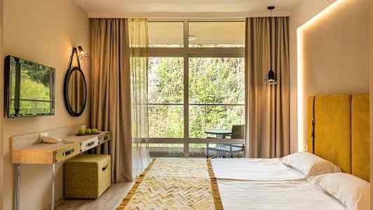 Et hotellrom med en dobbeltseng, gul sengegavl, lite skrivebord, og en stor vindusfront mot naturen. (AI-generert tekst)