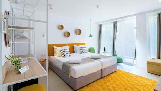 Et lyst og moderne hotellrom med to enkeltsenger, gule detaljer og store vinduer med gardiner. (AI-generert tekst)