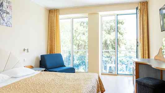 Rom med seng, blå lenestol og skrivebord, med store vinduer mot balkong. (AI-generert tekst)