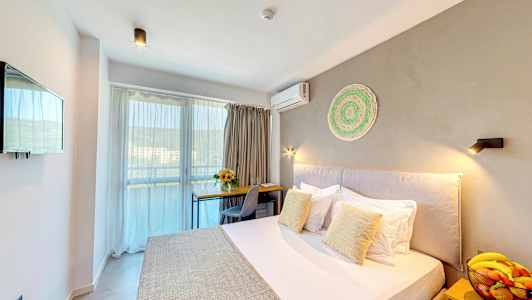 Et hotellrom med en seng, et skrivebord, en stol, en bolle med frukt på et nattbord og en balkong i bakgrunnen. (AI-generert tekst)