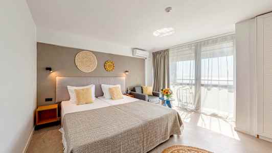 Et lyst hotellrom med en stor seng, grå sofa og balkong med utsikt. (AI-generert tekst)