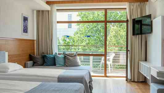 Et hotellrom med to senger, en sofa med fargerike puter og en glassdør som fører til en balkong. (AI-generert tekst)