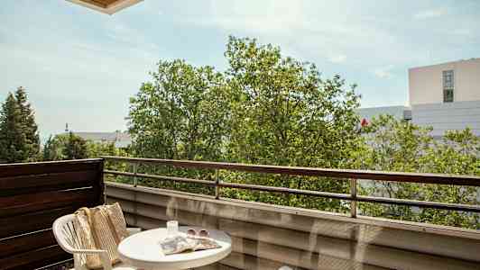 En balkong med et bord, to plaststoler, en veske, solbriller og en avis, samt utsikt over trær og bygninger. (AI-generert tekst)