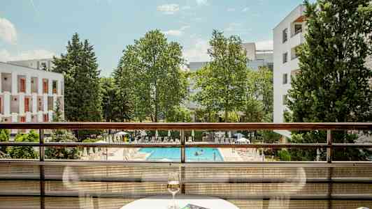 Utsikt fra en balkong med utsikt over et basseng og grøntarealer, inkludert et bord med et glass vin og en bok. (AI-generert tekst)