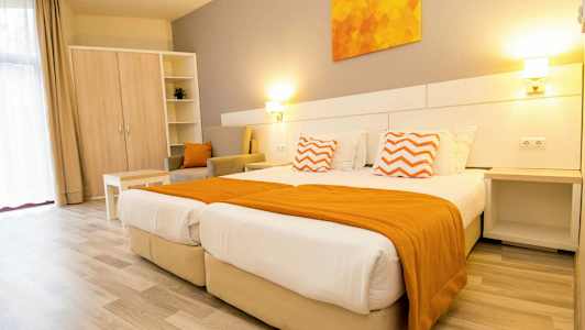 Et lyst hotellrom med to senger, orange detaljer og møbler. (AI-generert tekst)