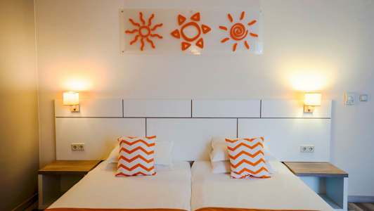 Et hotellrom med to enkeltsenger, oransje puter og veggdekorasjoner i form av soler. (AI-generert tekst)